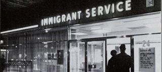 Svartvit kvällsbild på entrén till Immigrant service med en lysande neonskylt 