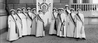 Gruppfoto med kvinnor klädda i vita, fotsida klänningar, studentmössor och ordensband, hållande ett standar