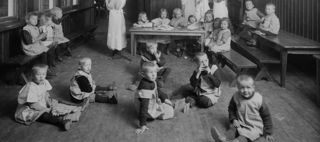 Svartvitt foto av barn, vissa sittande på golvet, samt kvinnor och flickor i förkläden och hucklen. 