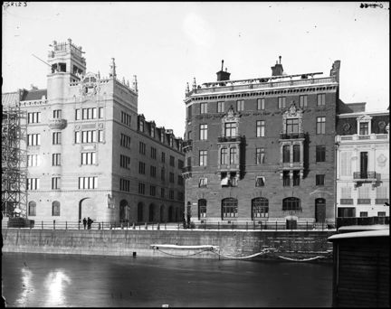 I förgrunden ett vattendrag och i bakgrunden två flervåningshus med utsirade fasader.
