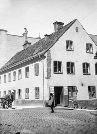 Vid en gata belagd med gatsten ligger ett 2,5 våningar högt stenhus. I bottenvåningen ligger en mekanisk verkstad, som har skyltar i fönster och på dörrparet. På fasaden görs reklam ved och kol. En flicka går på gatan och en bit bort kommer en häst med vagn.