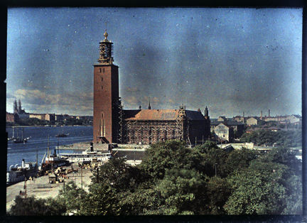 Färgbild med Stockholms stadshus i centrum. Byggställningar syns fortfarande runt huset. I nedre delen av bilden syns grönskande träd och till vänster Mälarens vatten.