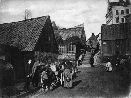 Svart-vit bild med människor längs en gata. En kvinna i mitten av bild bär kjol och sjal över huvudet, och håller i en kanna. I bakgrunden syns en trappa, flera trähus, ett högt stenhus och telefonledningar.
