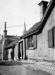 Två barn står vid porten till Iversons Gränd 5, nu Iversonsgatan. Pojken håller i ett tunnband. I fonden västerut ses tornet på Johannes kyrka