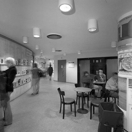 Biblioteksbesökare, några bord och stolar, vägg med tidskrifter.