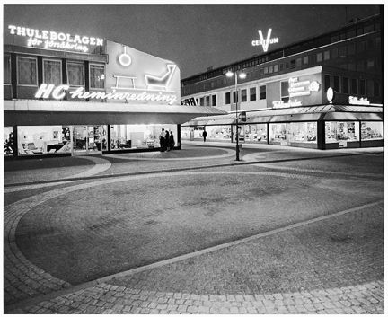 Vällingbyplan med neonskyltar och gatubelysning, människor tittar i skyltfönster