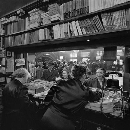 Genom en butikslucka betjänar två äldre kvinnor människor som ska köpa tidningar, böcker eller vykort. Utanför luckan är det en stor väntsal.