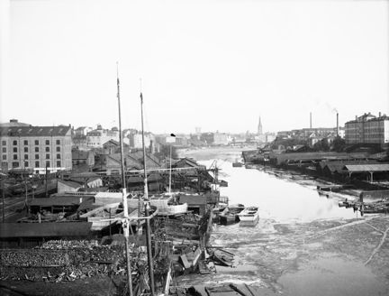 En kanal med bebyggelse på båda sidor. Längst fram master från en båt.
