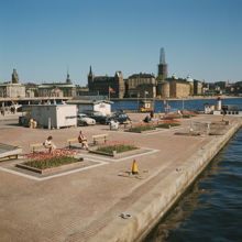 Klara Mälarstrand (""Stadshuskajen"") med bänkar och planteringar. Kontor för Forsman Sightseeing och Pressbyråkiosk. I fonden Riddarholmen