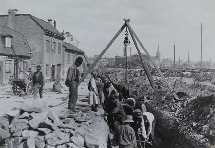 Flera män i hattar i ett gravt dike, en förman tittar ner på dem. Klara kyrka i fonden.