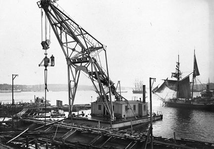 En gammal lyftkran ligger på en pråm i vattnet. Bakom syns flera segelfaryg.