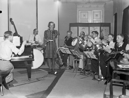Alice Babs stående vid en mikrofon och sittande musiker på vardera sida.