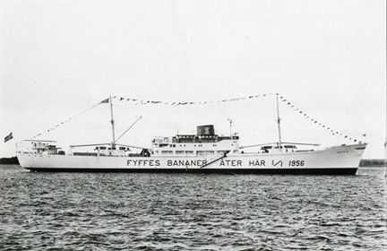 Kylfartyget "San Blas" ligger i vattnet, dekorerat med flaggspel längs hela däck. På skrovsidan står texten ”FYFFES BANANER ÅTER HÄR 1956”. Fartyget är vitt och har en mörk skorsten med rederiets märke.