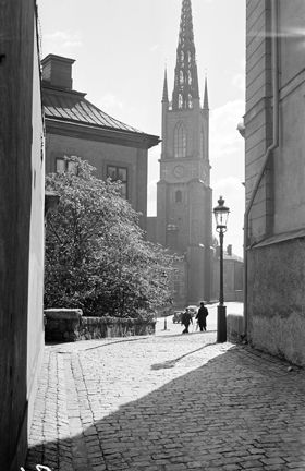 Schering Rosenhanes gränd öppnar sig mot Birger Jarls torg. Ett par personer och bilar skymtar. Riddarholmskyrkans torn syns i bakgrunden.