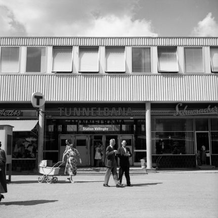 Människor rör sig utanför tunnelbanenedgången, bland andra en kvinna med barnvagn. En telefonkiosk skymtar.