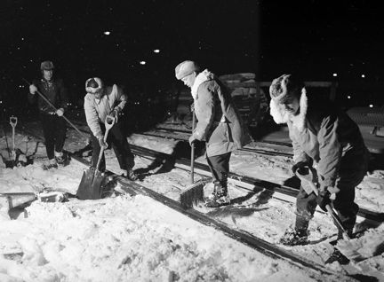 Fyra män står på ett spår och skottar snö med skovlar. De står i ett ljust område men runtomkring dem är det mörkt. Männen har tjocka jackor med pälskragar och vintermössor med uppfällda öronlappar.