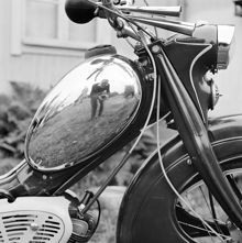 Närbild av moped med kromad bensintank. Fotografen speglad i tanken