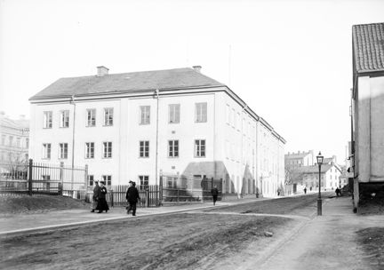 Intill en gata, utan gatubeläggning och ganska lerig, ligger ett stort vitt hus i tre våningar. Runt husets tomt går ett lite högre staket. Solen skiner och några människor promenerar längs gatan. 