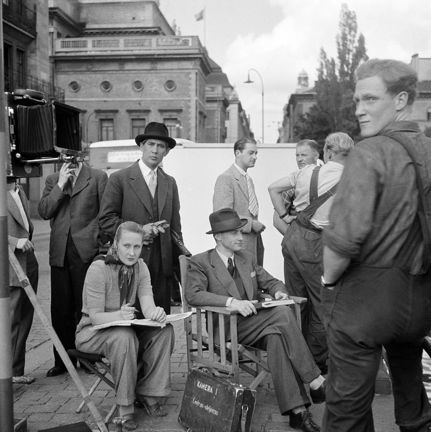 Filminspelning. Regissör och andra medarbetare samt skådespelare sitter och står och väntar på nästa scen.