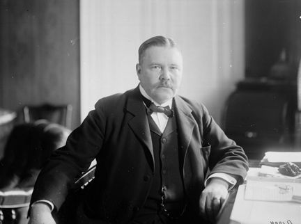 En man i kostym, fluga och mustasch sitter vid sitt skrivbord. Han tittar in i kameran.