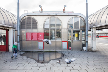 Gullmarsplans busshållplats, byggnad med fönster och dörr i plåt. Duvor flyger iväg, ett barn går förbi