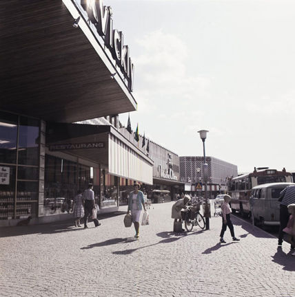 Trottaren utanför Farsta centrum. Människor promenerar och bredvid står en bil och en buss parkerad.