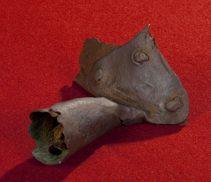 Medeltida grytskaft av metall med tre nitar. Fotograferad mot röd bakgrund. 