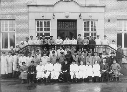 Gruppbild av personalen vid Slakthuset i Johanneshov år 1936. Ett femtiotal personer, både män och kvinnor,  sitter eller står framför en byggnad i ljust tegel. Några personer står på en trappa med metallräcke.