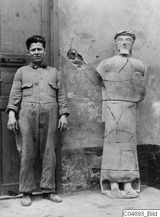En man, Toulis Souidos, i arbetskläder intill en terracottafigurin som han lagat. Skulpturen är lika stor som han och man ser tydligt sprickorna i godset.