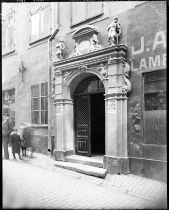 En port med dubbeldörrar, omsluten av en stenportal med skulptural utsmyckning. Utanför på gatan står flera barn och tittar åt fotografens håll. Den ena dörren står öppen.